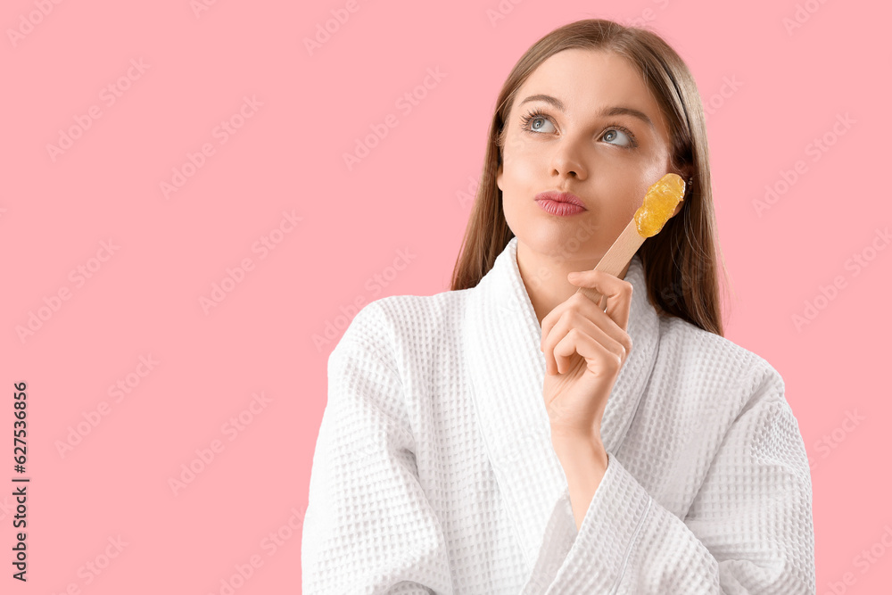 Thoughtful young woman holding spatula with sugaring paste on pink background