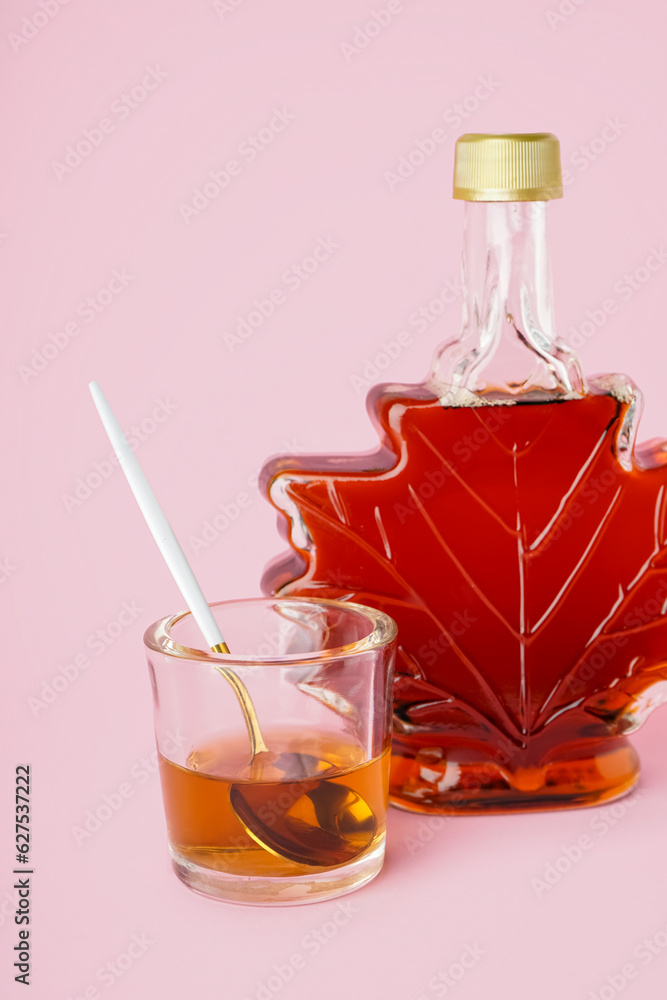 Bottle and spoon in glass of tasty maple syrup on pink background