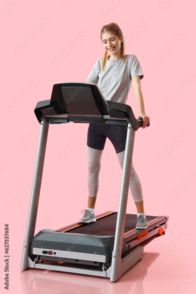 Sporty young woman training on treadmill against pink background