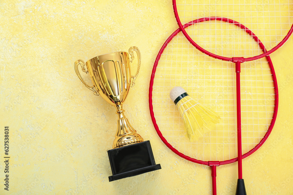 Gold cup with badminton rackets and shuttlecock on yellow background