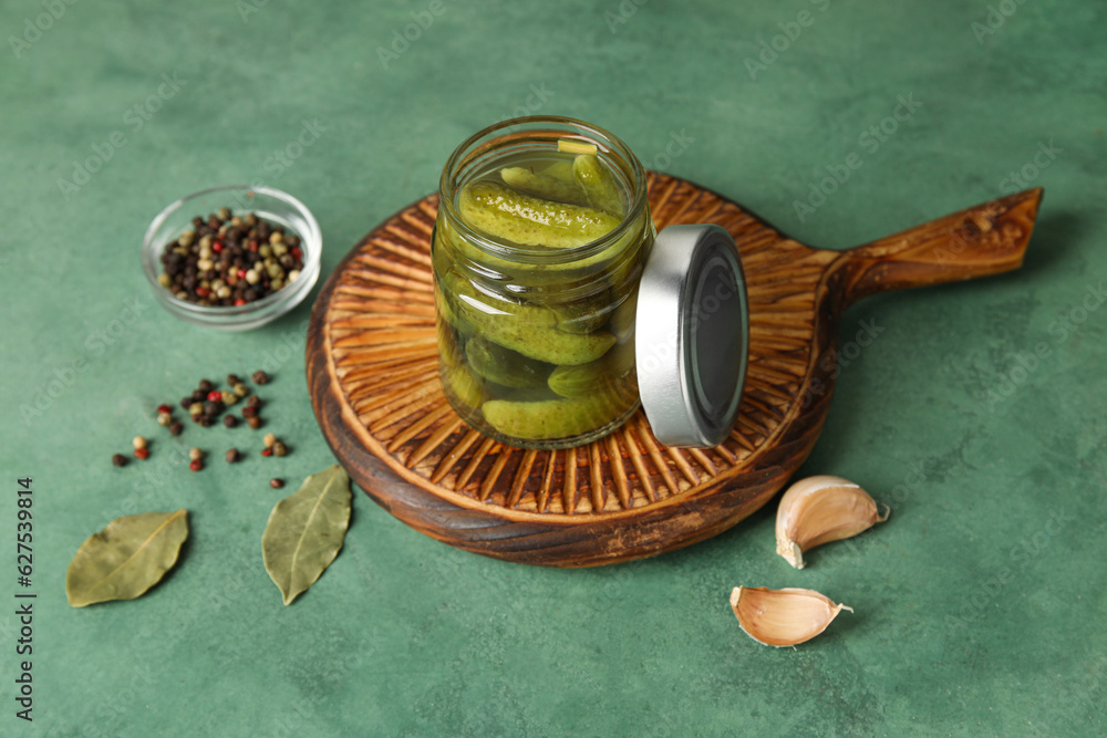 Jar with tasty fermented cucumbers, wooden board and ingredients on green background