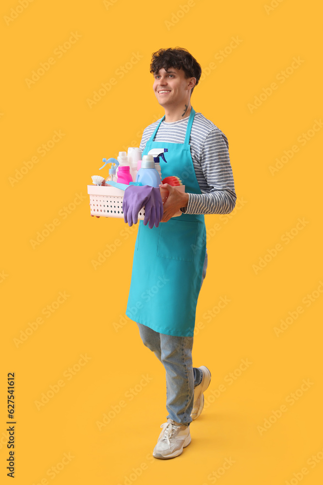 Young man with basket of cleaning supplies on yellow background