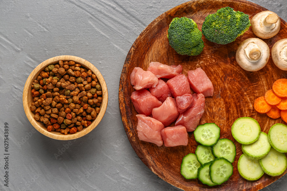 Bowl with dry pet food, raw meat and natural products on color wooden background