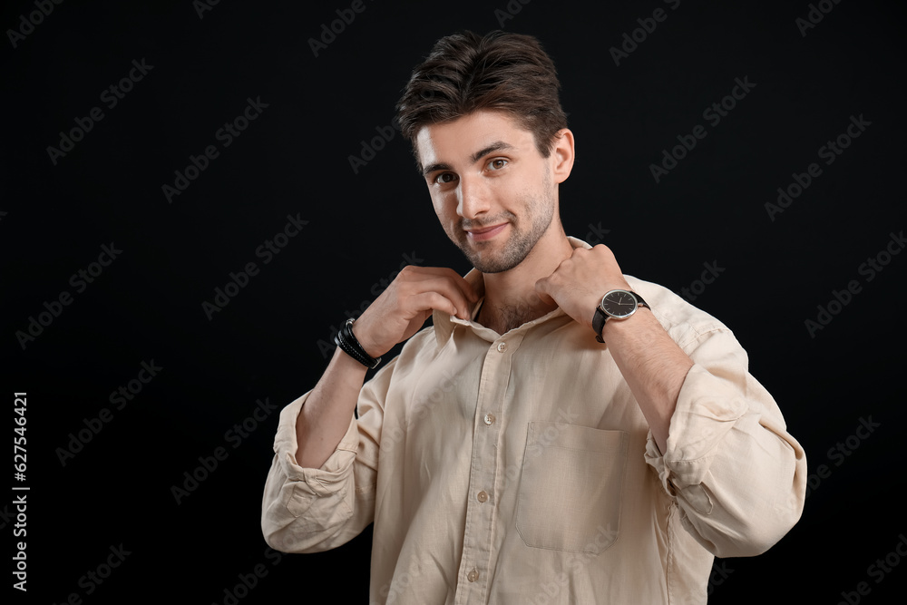 Stylish young man with wristwatch adjusting shirt on black background