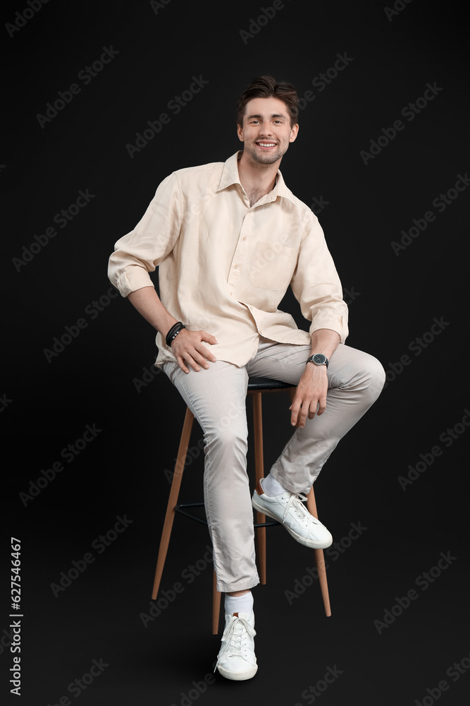 Handsome happy young man with wristwatch sitting on chair against black background