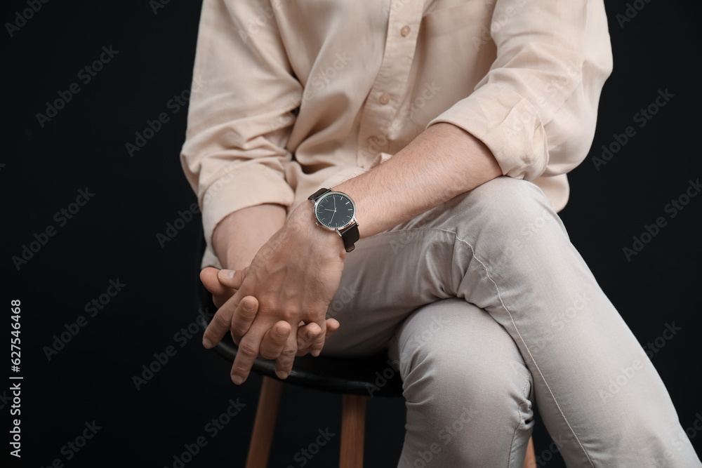 Stylish handsome young man with wristwatch sitting on chair against black background, closeup