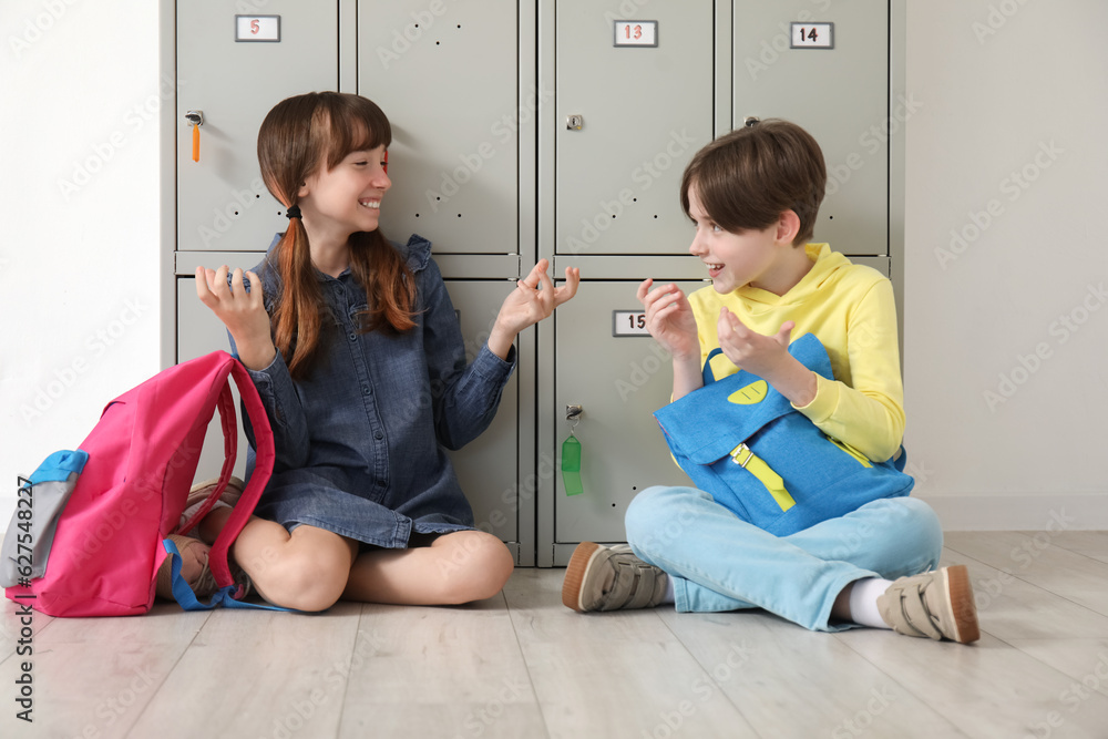 Little pupils with backpacks sitting near locker at school