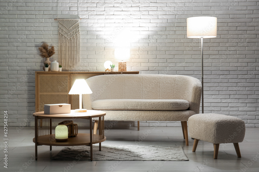 Interior of dark living room with cozy white sofa, pouf, coffee table and glowing lamps