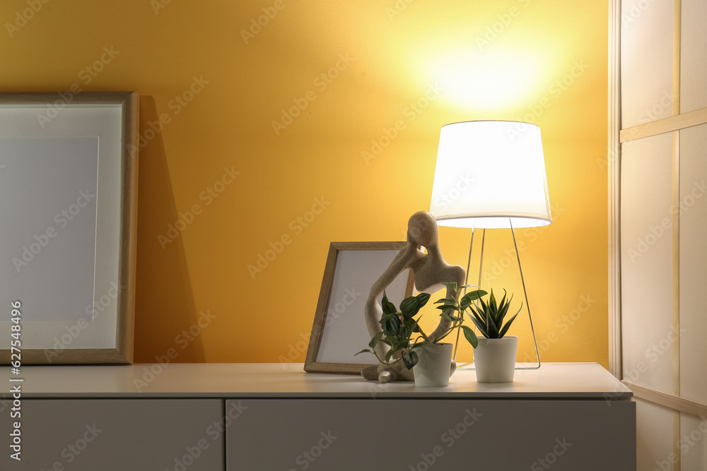 Glowing lamp, blank frames, houseplants and decorative figurine on cabinet in dark room