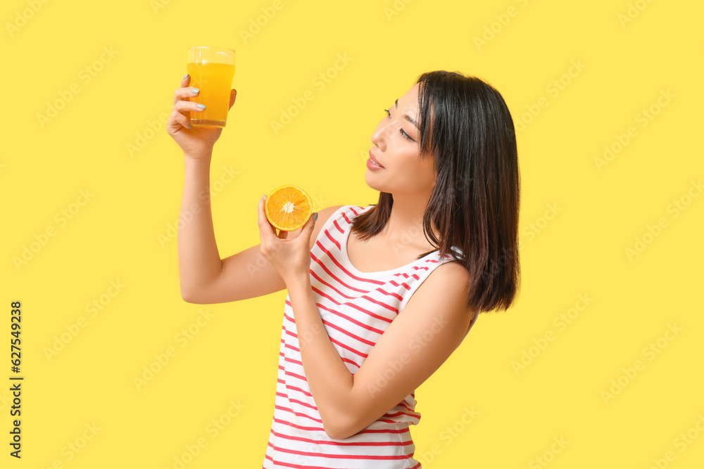 Beautiful Asian woman with orange and glass of juice on yellow background