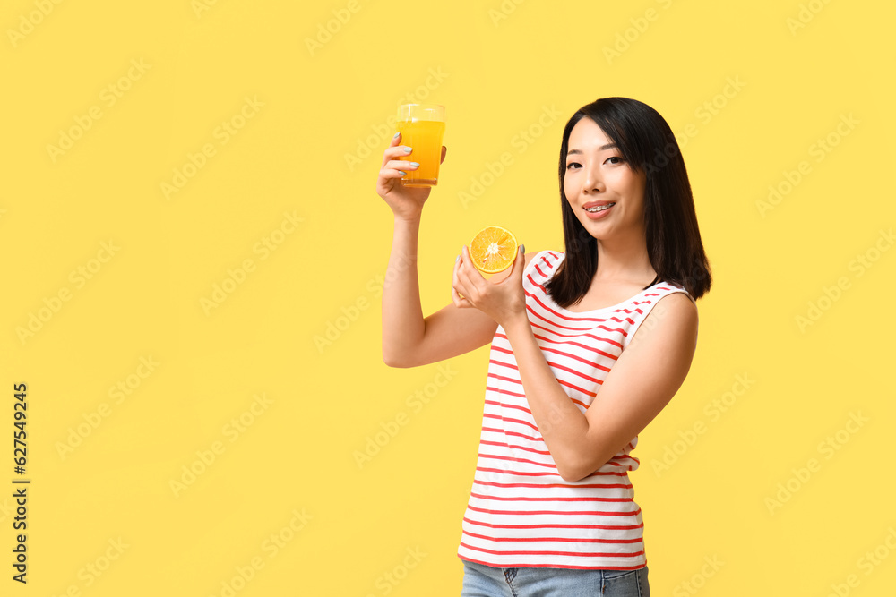 Beautiful Asian woman with orange and glass of juice on yellow background