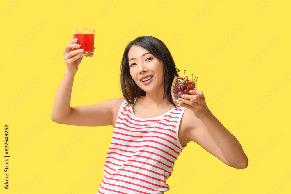 Beautiful Asian woman with cherries and glass of juice on yellow background