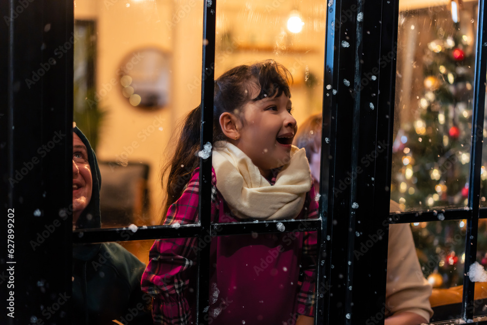 Adorable child looking at the window and first snow flakes with family. 