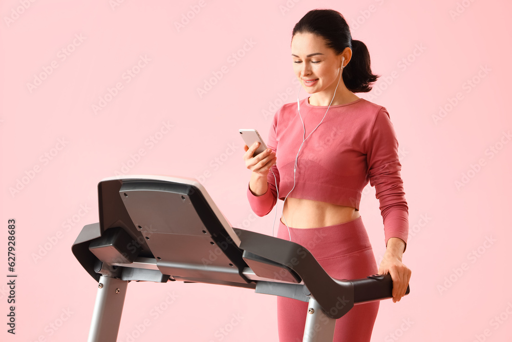 Beautiful woman with mobile phone and earphones training on treadmill against pink background