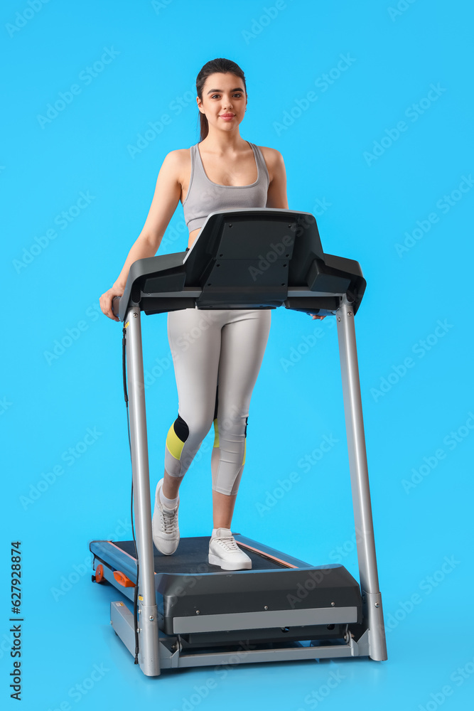 Sporty young woman training on treadmill against blue background