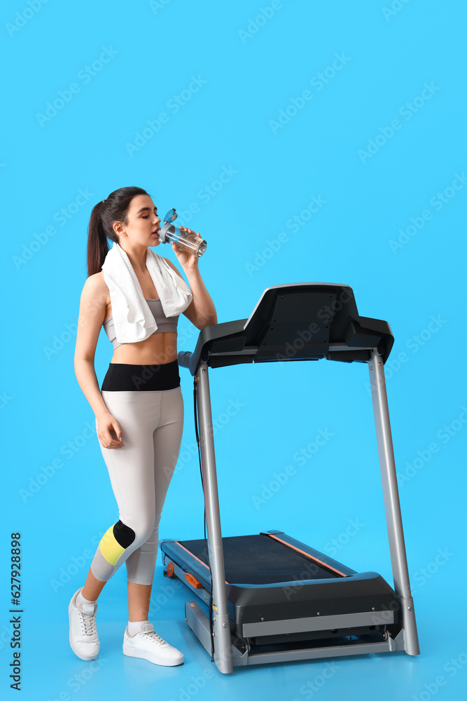 Sporty young woman with treadmill drinking water on blue background