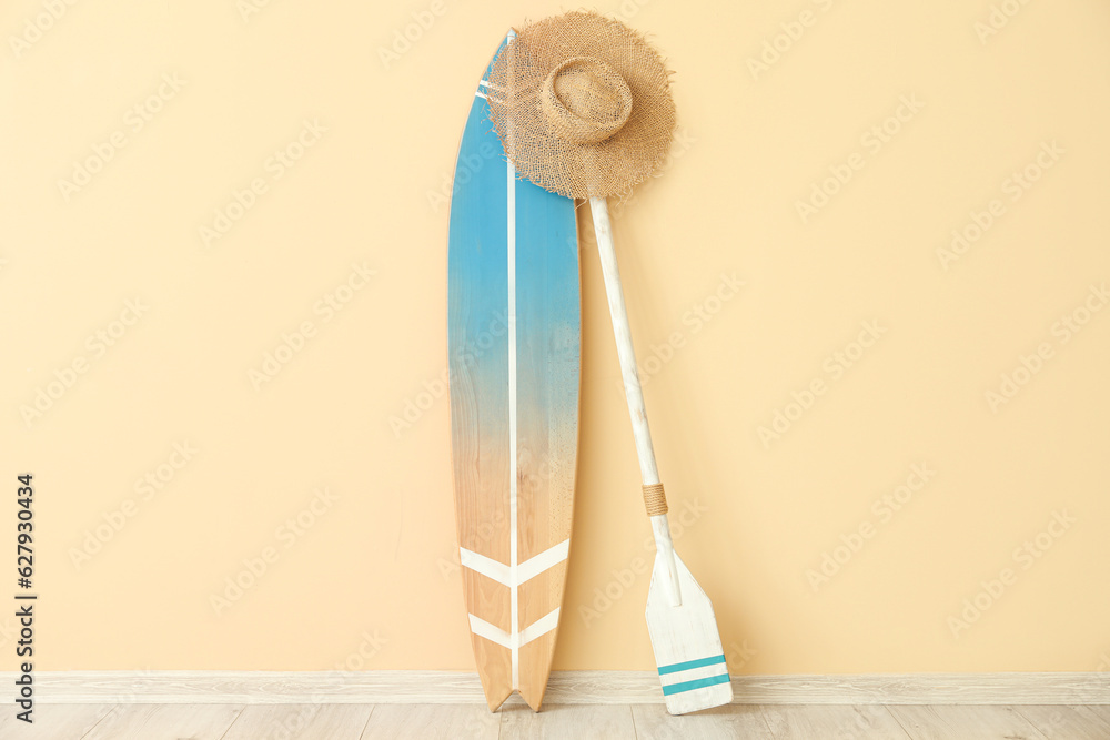 Wooden surfboard with paddle and hat near beige wall