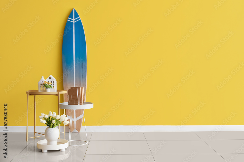 Tables with flowers, books and surfboard near color wall in room