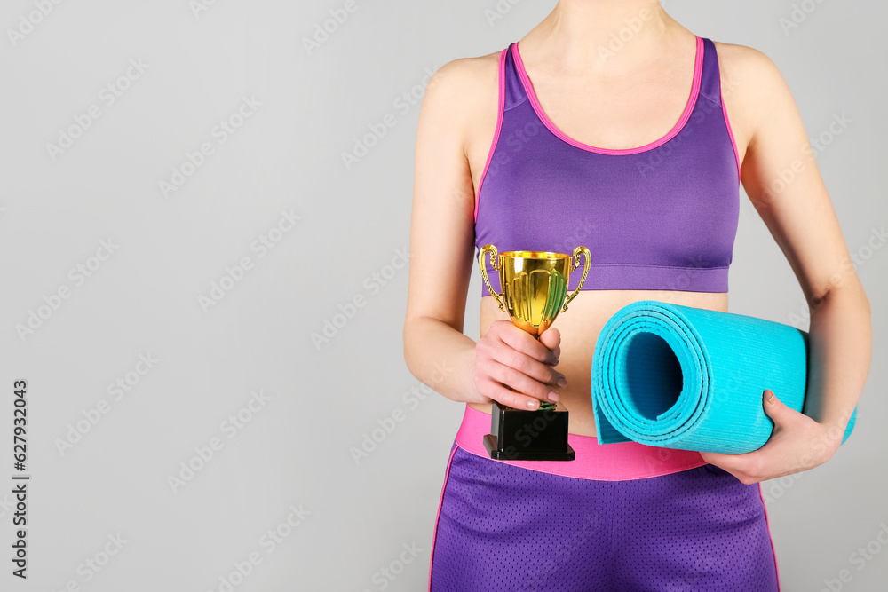 Sporty young woman with fitness mat and gold cup on grey background