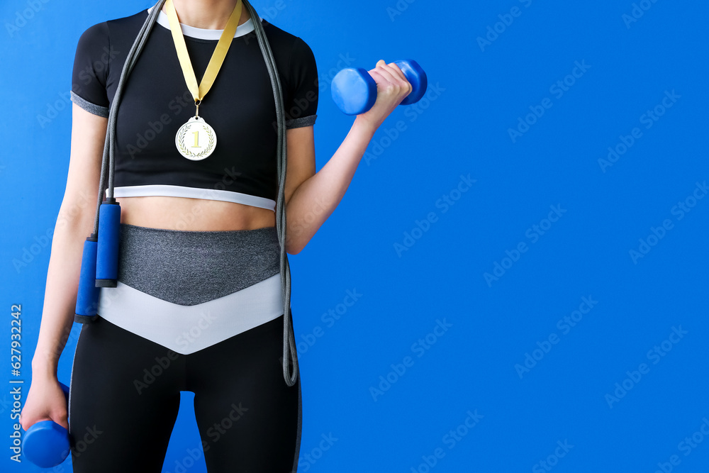 Sporty young woman with first place medal, skipping rope and dumbbells on blue background