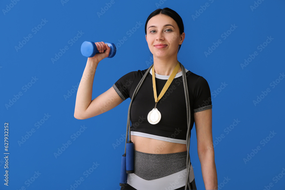 Sporty young woman with first place medal, skipping rope and dumbbell on blue background