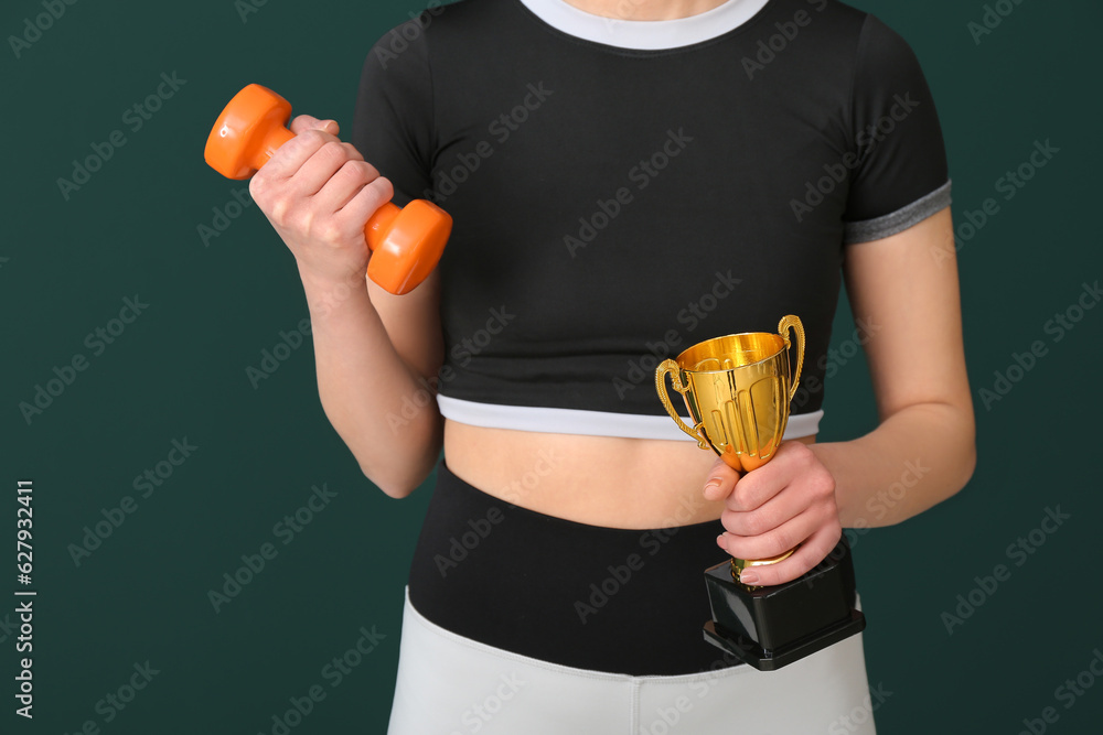 Sporty young woman with gold cup and dumbbell on green background, closeup