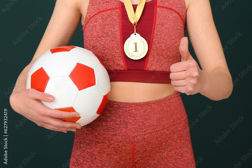 Female football player with first place medal and ball showing thumb-up on green background, closeup