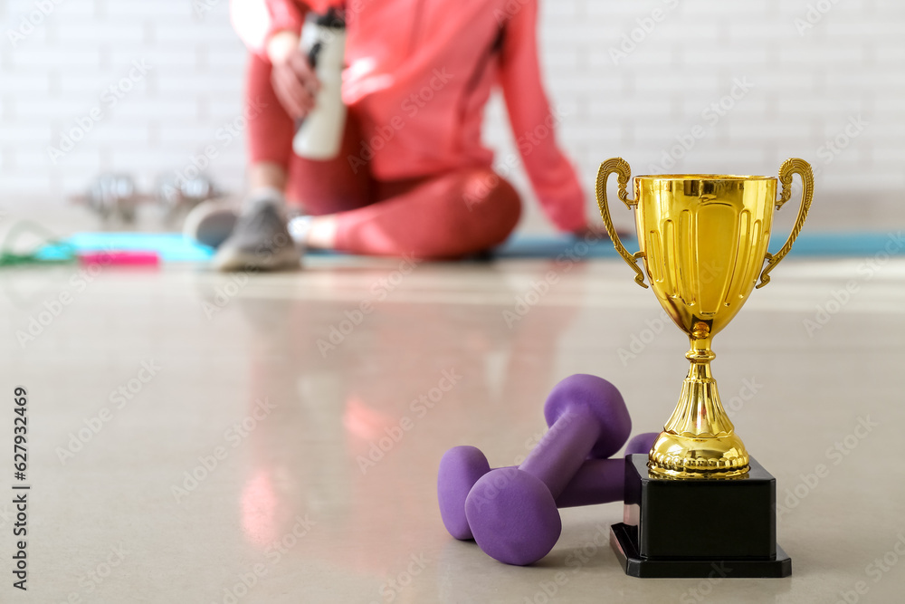 Gold cup with dumbbells on floor in gym, closeup