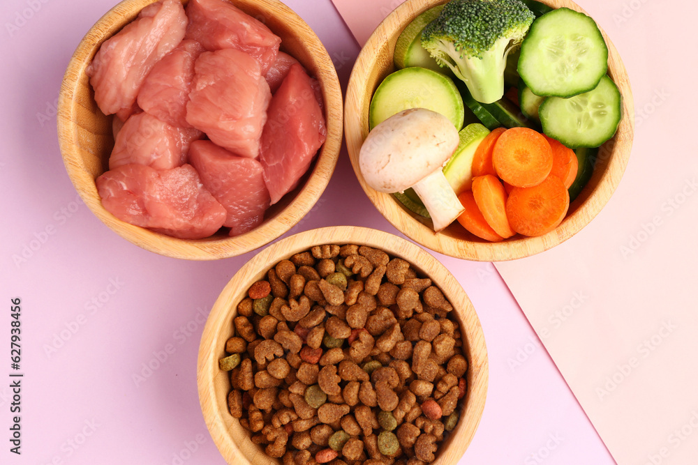 Bowls with dry pet food, raw meat and natural products on pink background, closeup