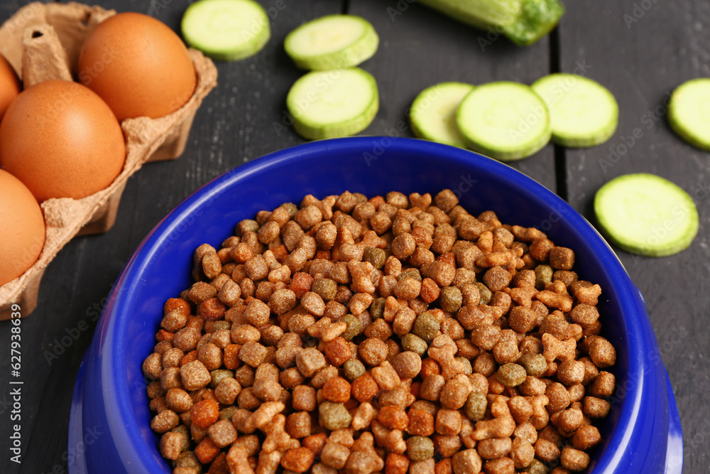 Bowl with dry pet food and natural products on dark wooden background, closeup