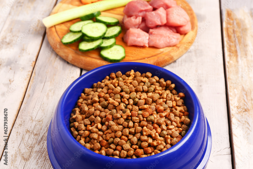 Bowl with dry pet food, raw meat and natural products on light wooden background, closeup