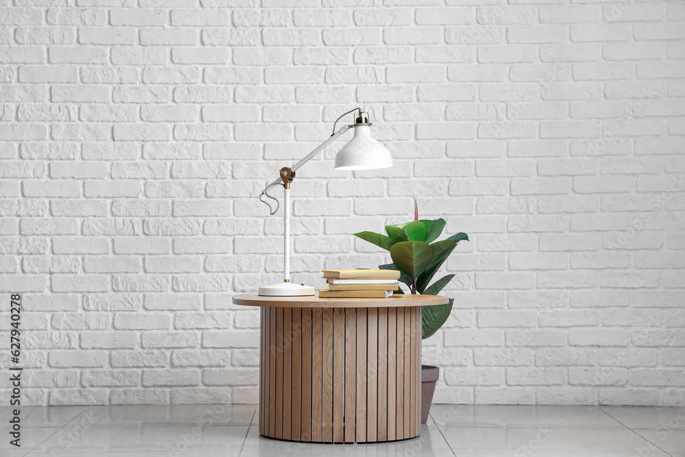 Table with glowing lamp, books and houseplant near white brick wall in room