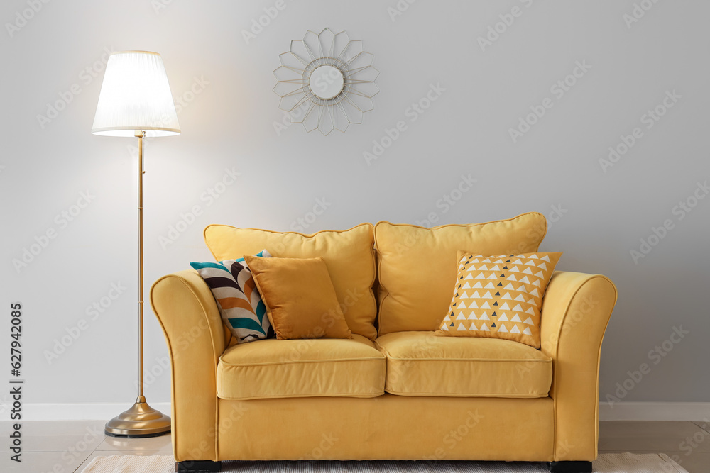 Glowing lamp, yellow sofa with cushions and mirror near white wall