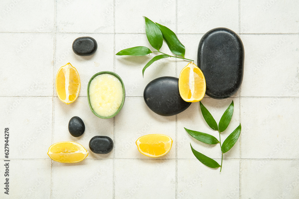 Bowl of lemon body scrub with spa stones and plant branches on white tile background