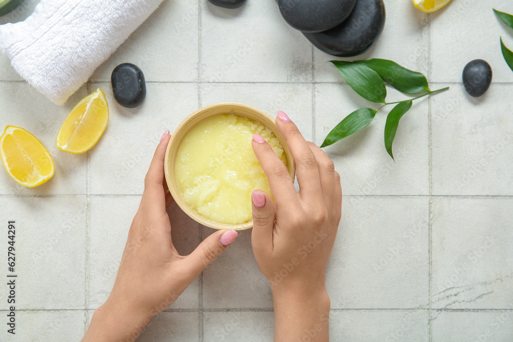 Female hands with bowl of lemon body scrub, spa stones and towel on white tile background