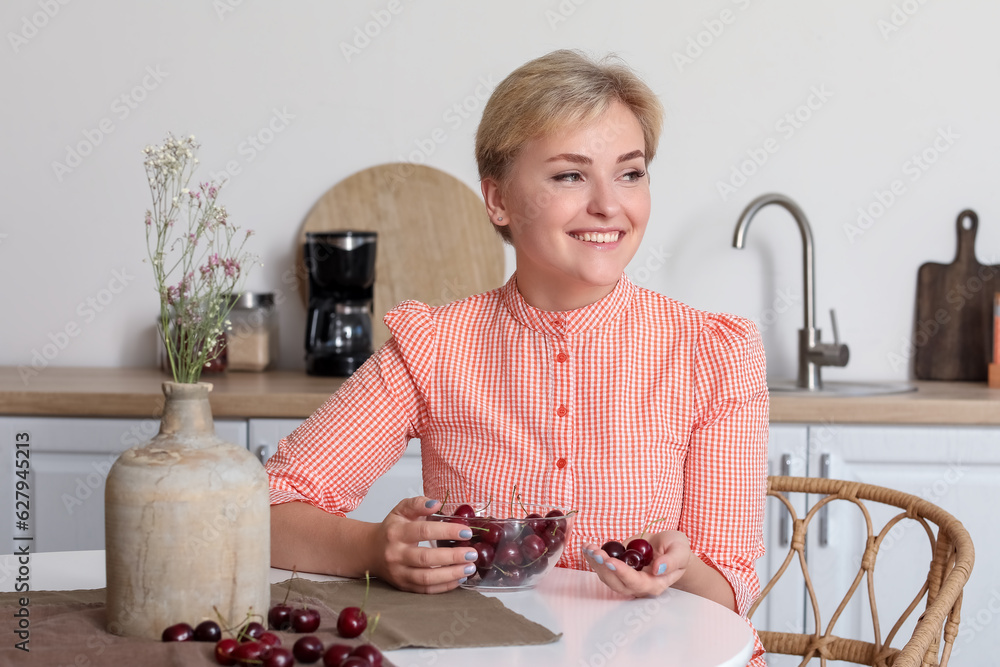 Beautiful young happy woman with bowl of ripe cherries sitting at table in kitchen