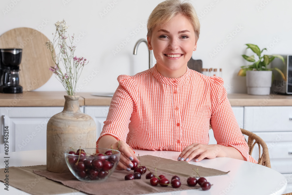 Beautiful young happy woman with bowl of ripe cherries sitting at table in kitchen