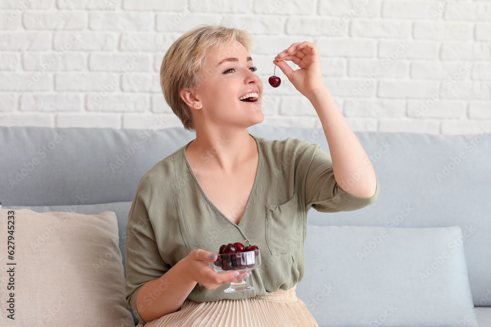 Beautiful happy young woman sitting on sofa in living room and eating ripe cherries