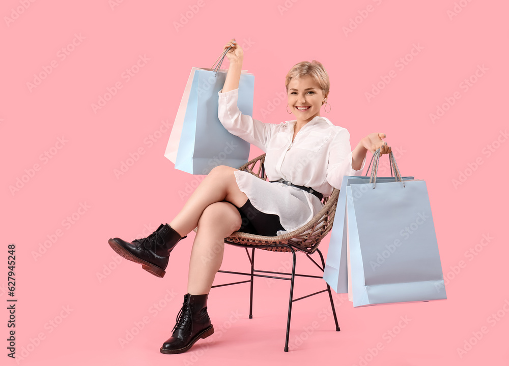 Beautiful young woman with shopping bags sitting on chair against pink background