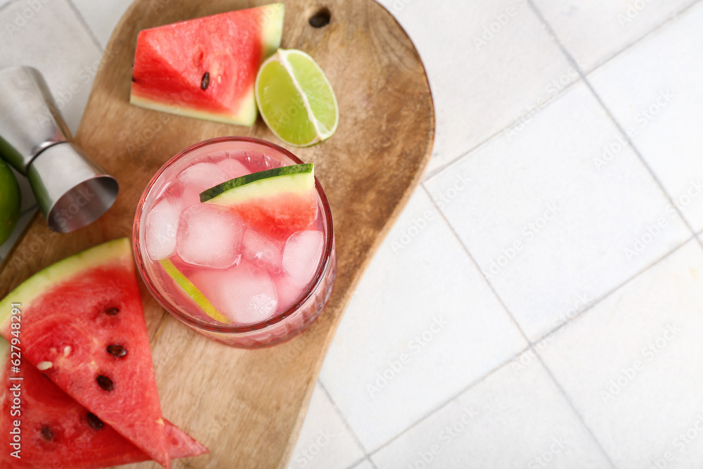 Board with glass of tasty watermelon cocktail, jigger and lime on white tile background