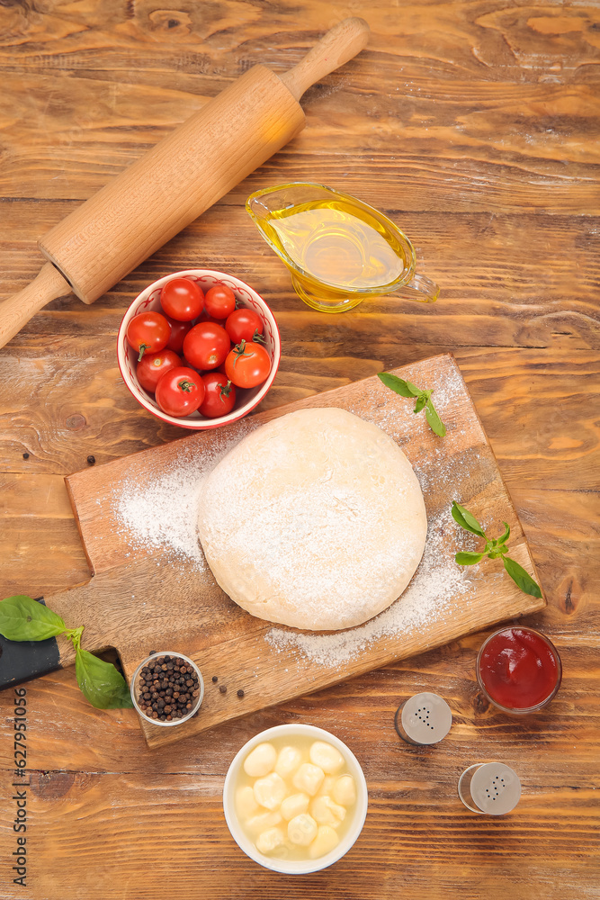 Board with raw dough and ingredients for preparing pizza on wooden background