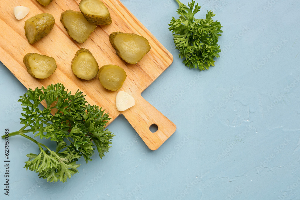 Wooden board with pickled cucumbers, garlic and parsley on blue background