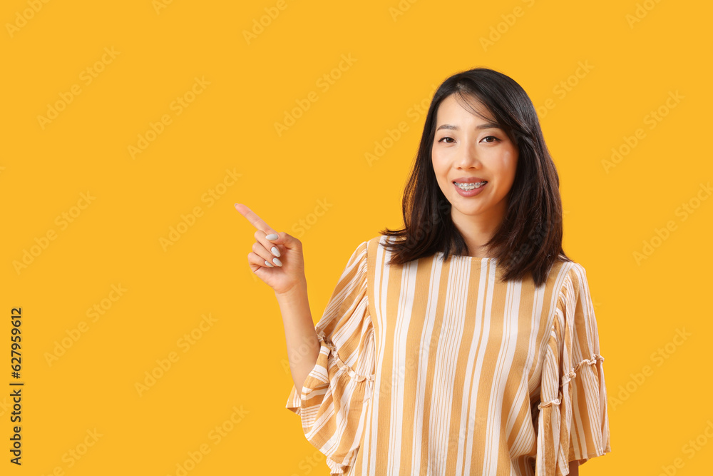 Beautiful Asian woman pointing at something on yellow background. Dialogue concept