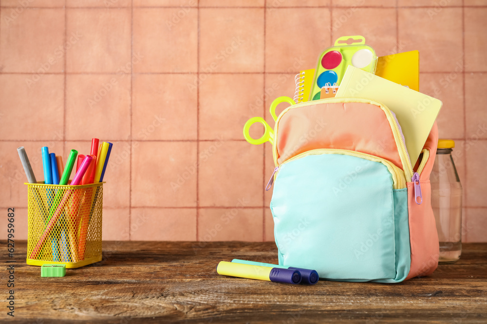 Colorful school backpack with notebooks, watercolors and cup of markers on wooden table near beige t