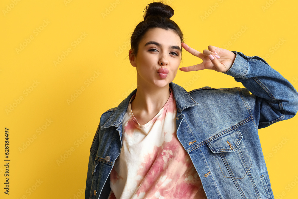 Young woman in tie-dye t-shirt showing victory gesture on yellow background