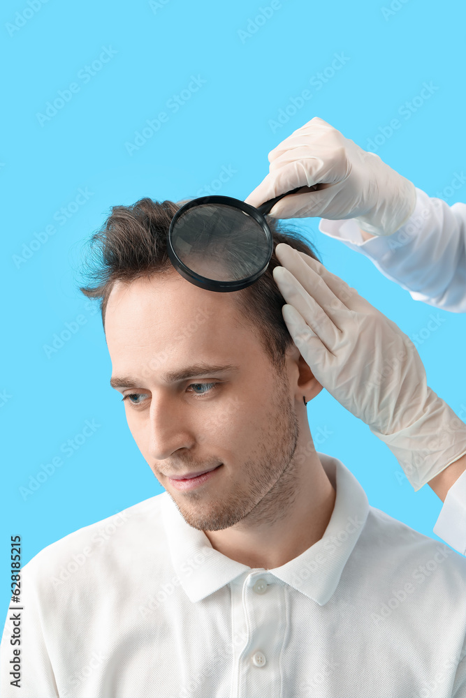 Doctor with magnifier examining young mans hair on blue background, closeup