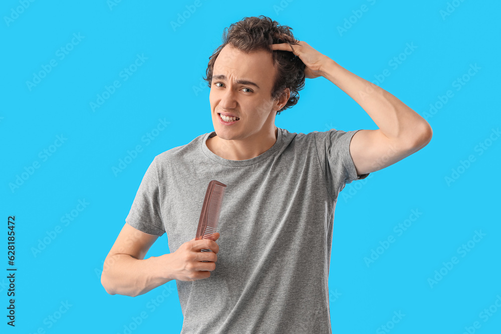 Upset young man with comb losing hair on blue background