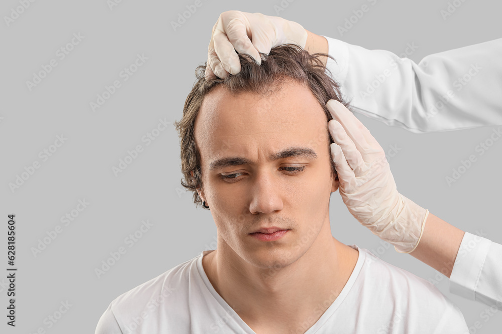 Doctor examining young man with hair loss problem on grey background, closeup