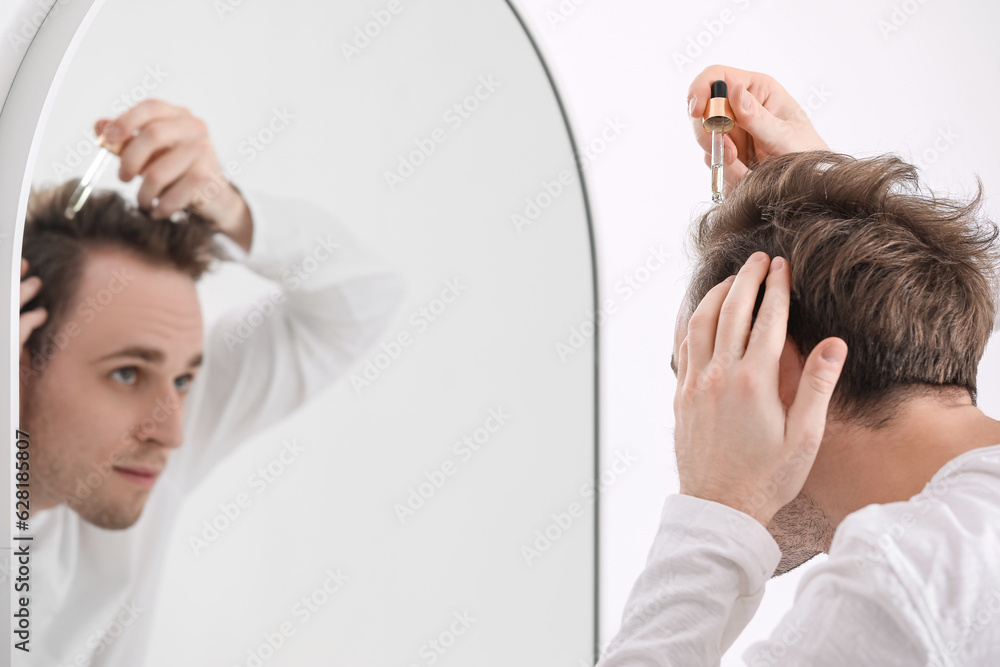 Young man using serum for hair growth near mirror in bathroom, closeup