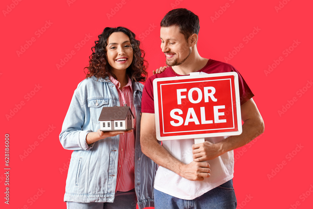 Young couple with FOR SALE sign and house model on red background